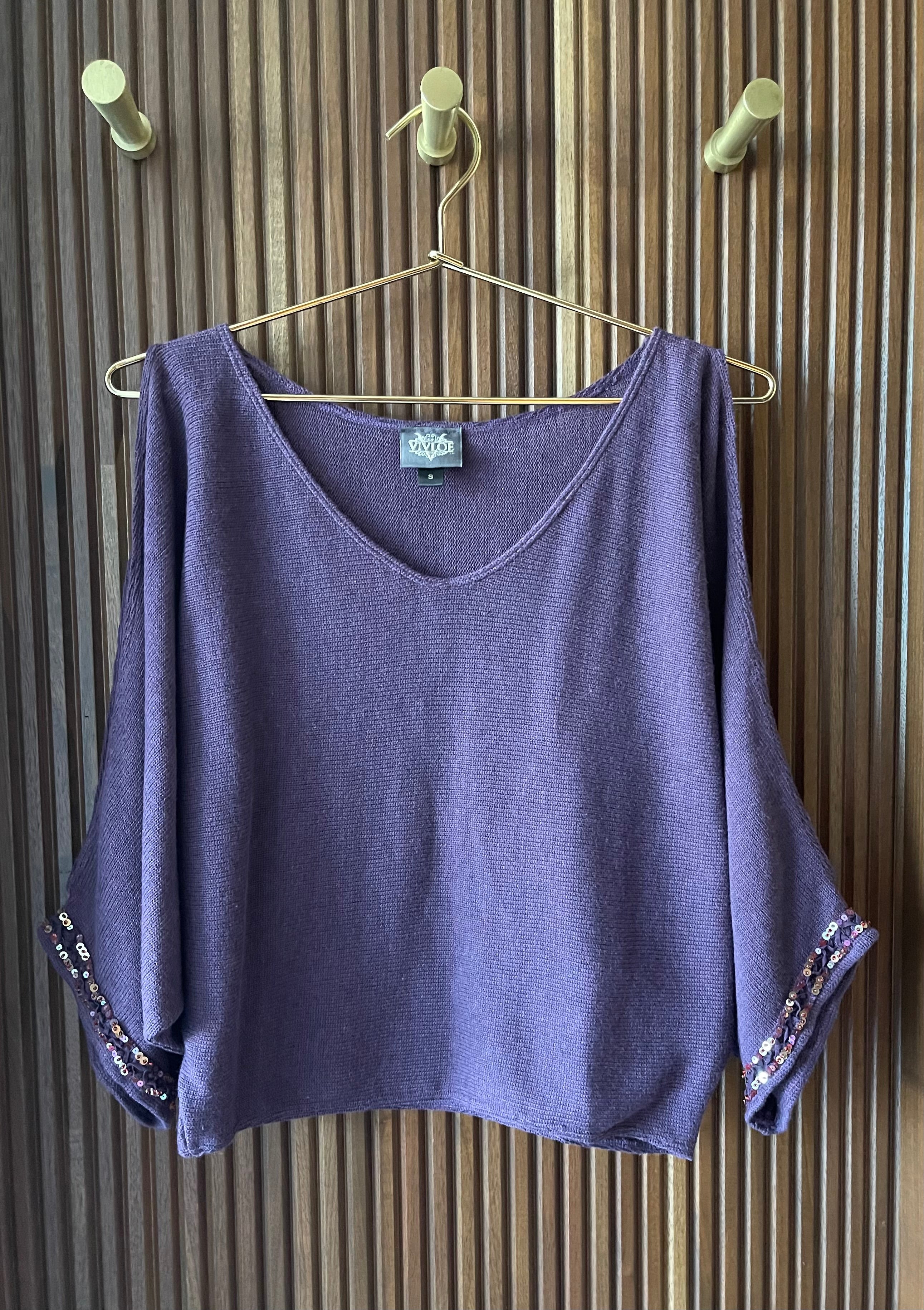 Pull over ladies sweater, jersey knit stitch, waist length with long sleeves with slit from shoulder to wrist, soft v-neck, hand beading on sleeve opening of vintage bugle, seed and sequin beads in pinks, purples and gold colors. The sweater is a deep blue purple, mulberry color. The sweater is hanging on a gold metal hanger against a textured wood wall.