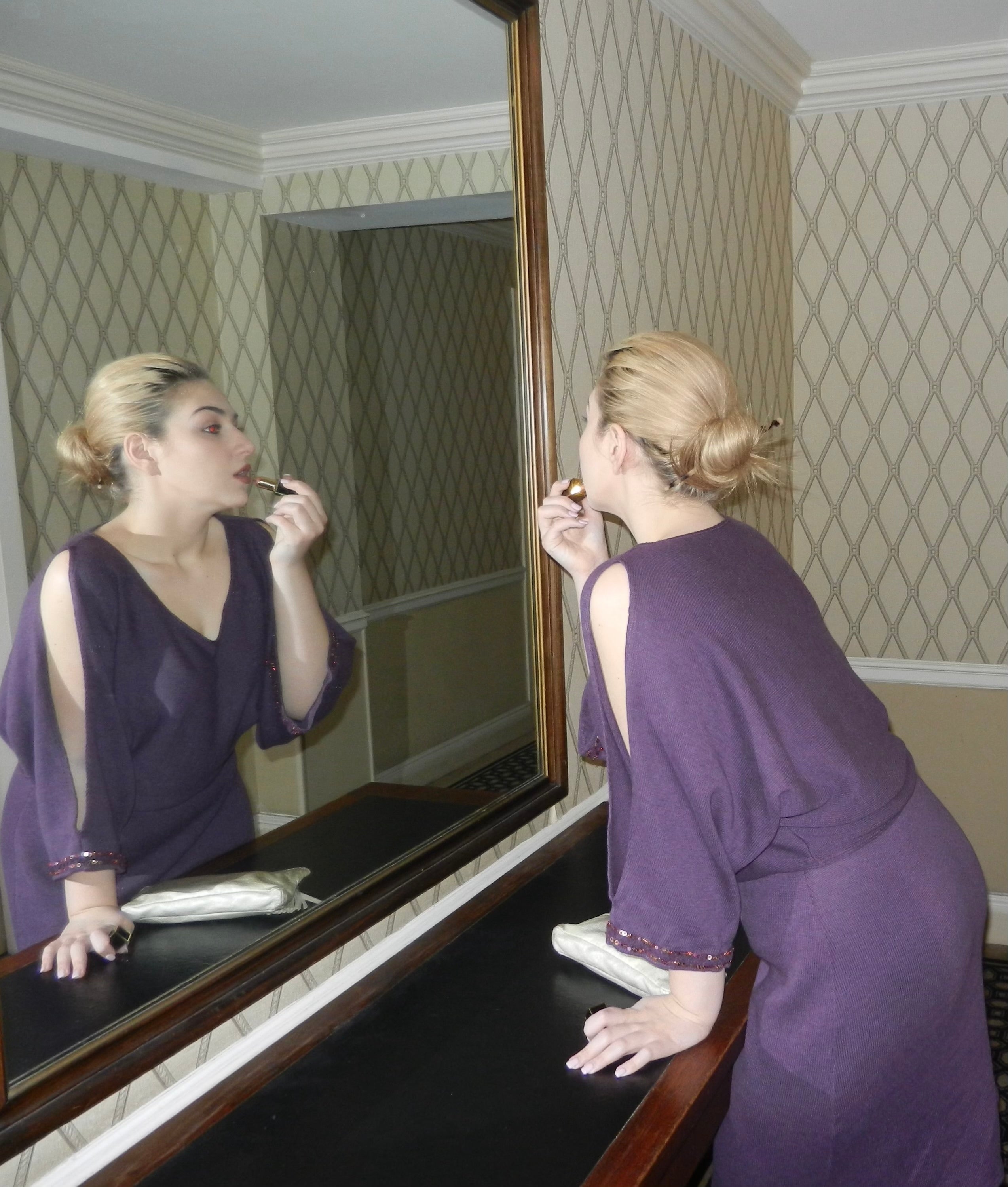 Woman applying lipstick in a mirror leaning on a console table in a hallway, wearing Vivloe's Spritz Sweater 2pc Dress in Mulberry color