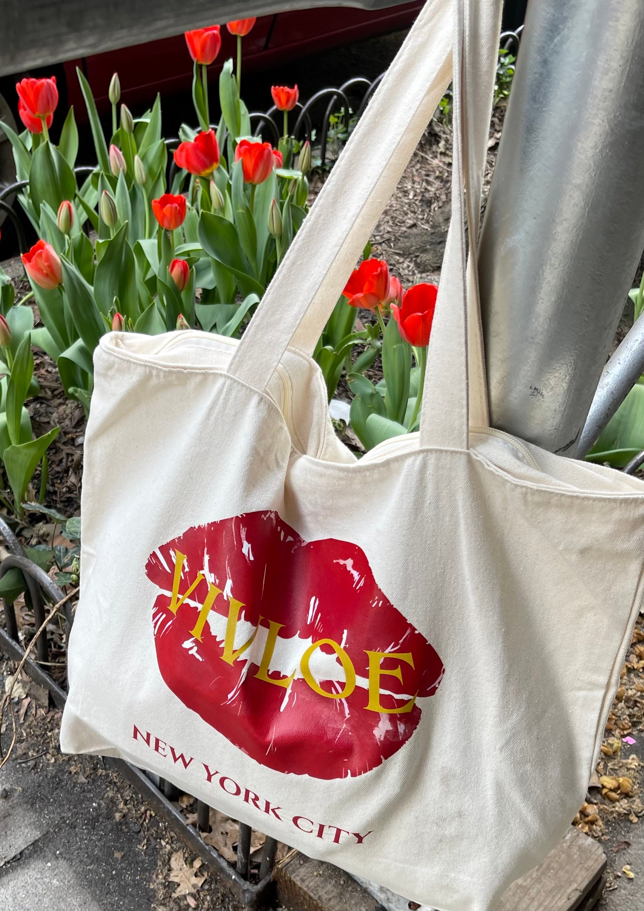Canvas natural color tote bag hanging from a pole next to a bed of red tulips on the sidewalk. Tote bag has a large red lip print with Vivloe written across in gold letters and New York City at the bottom in red.