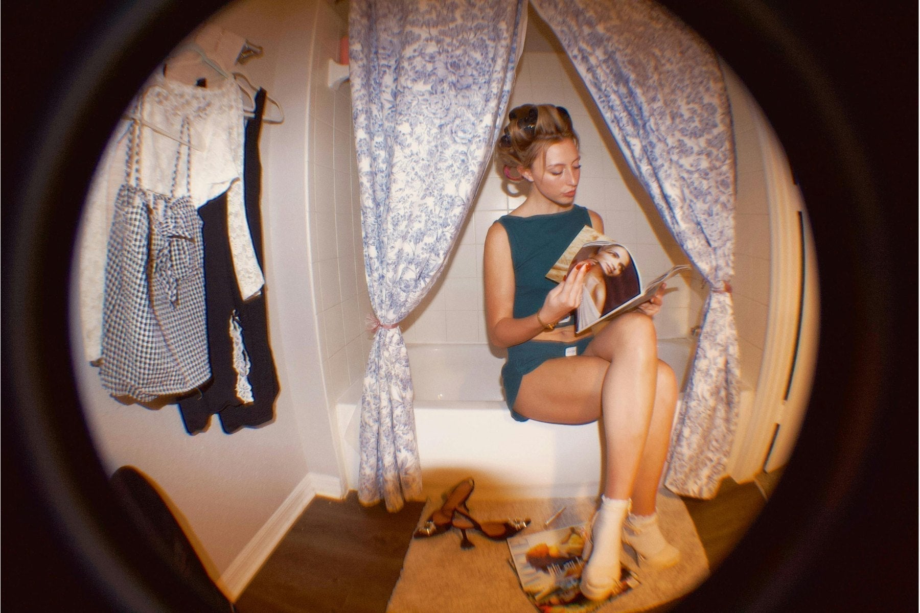 Image of woman sitting on bathtub edge reading a fashion magazine with sandals on the floor and another magazine on the floor, clothing hung on a hook on the wall next to her, viewed through a round hole in shadow