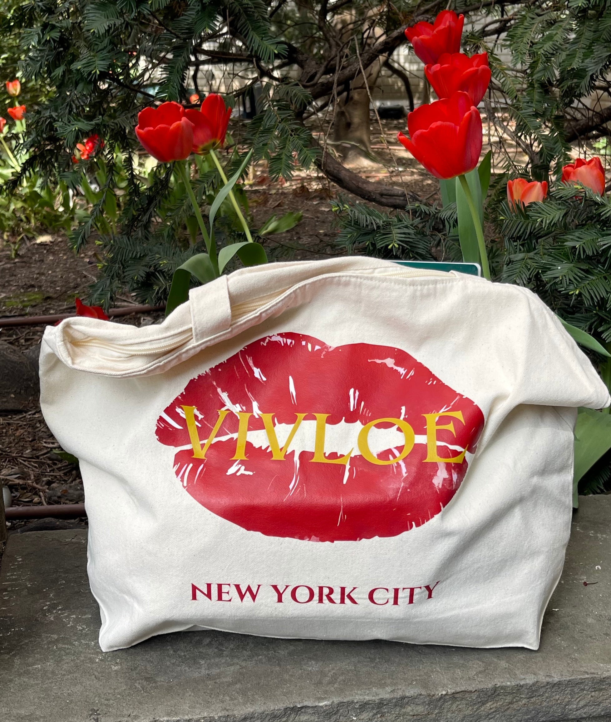 Natural off-white color canvas tote bag with red lip print and 'Vivloe New York City' text, placed outdoors with red tulips in the background.