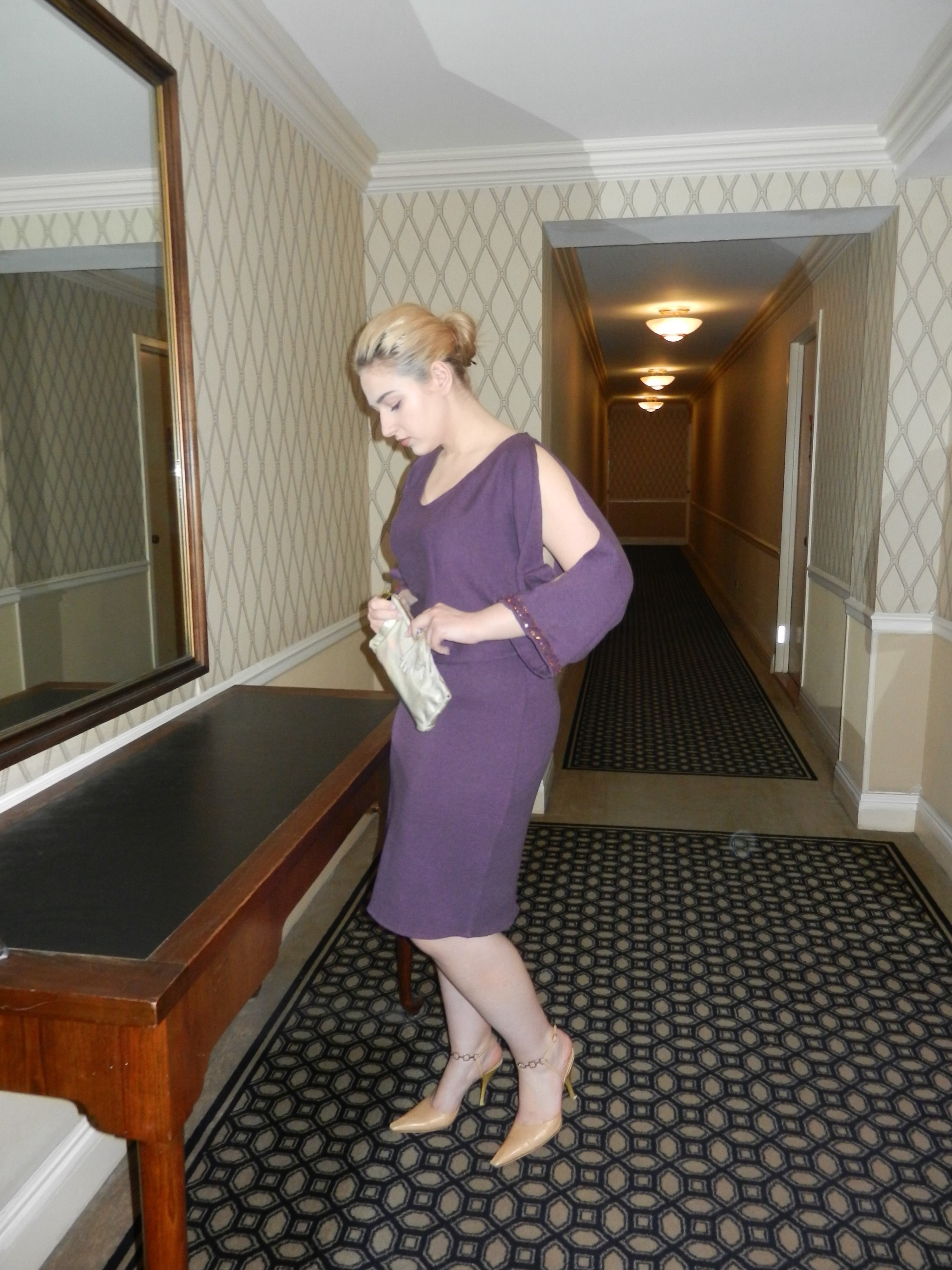 Woman in a purple dress standing in a hallway with patterned carpet and wallpaper.