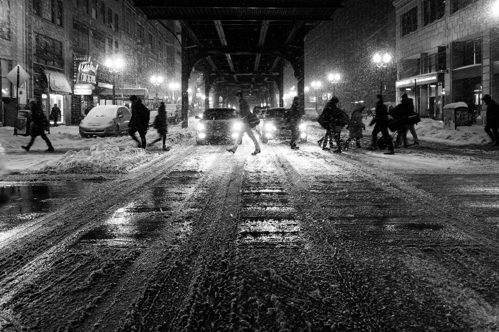 City street at night during a snowstorm with people and cars.